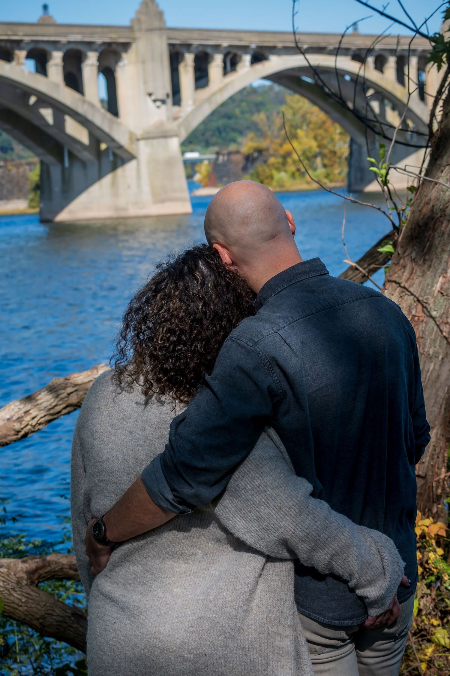 Couple embracing by a river with an arched stone bridge in the background on a sunny autumn day.