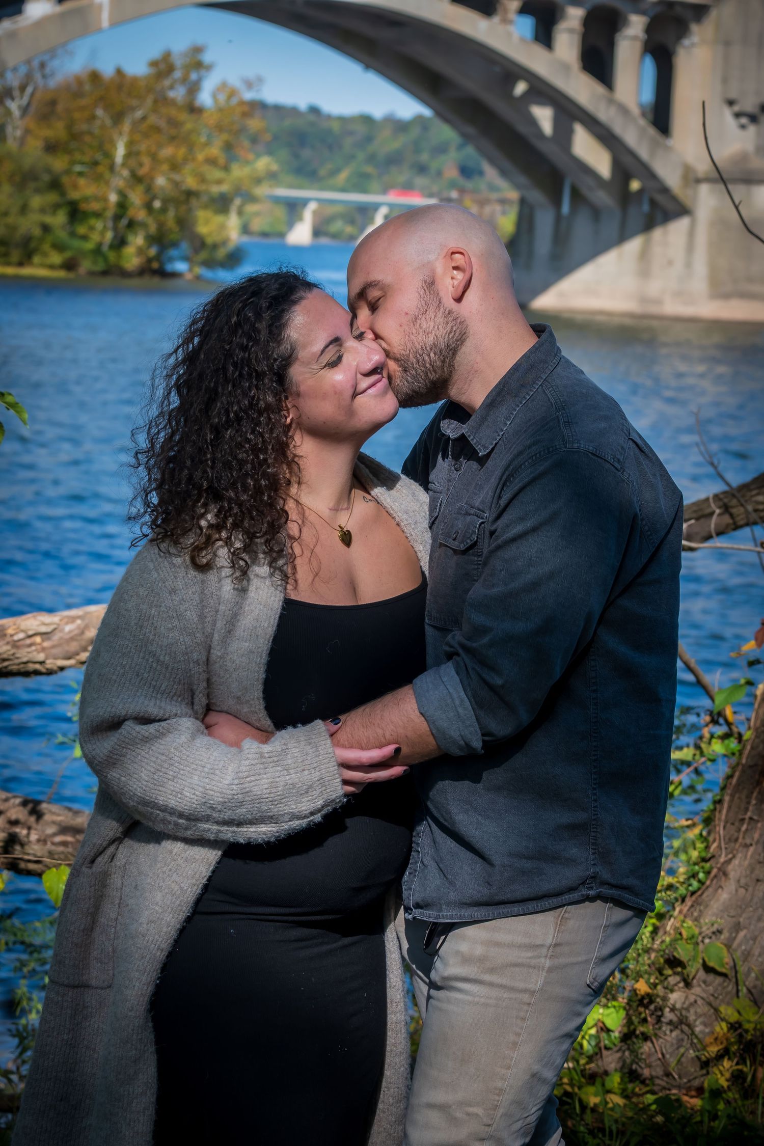 Couple sharing a tender kiss by a river with an arched bridge backdrop on a sunny autumn day.