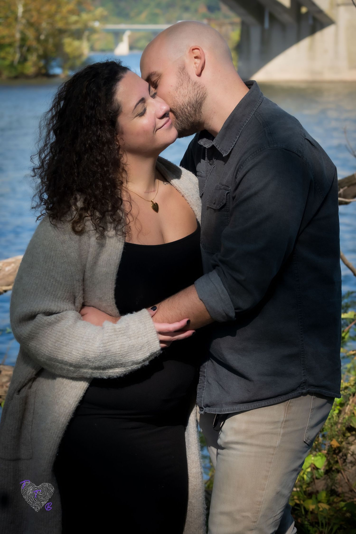 Couple sharing a tender moment near a river with a bridge in the background on a sunny day.