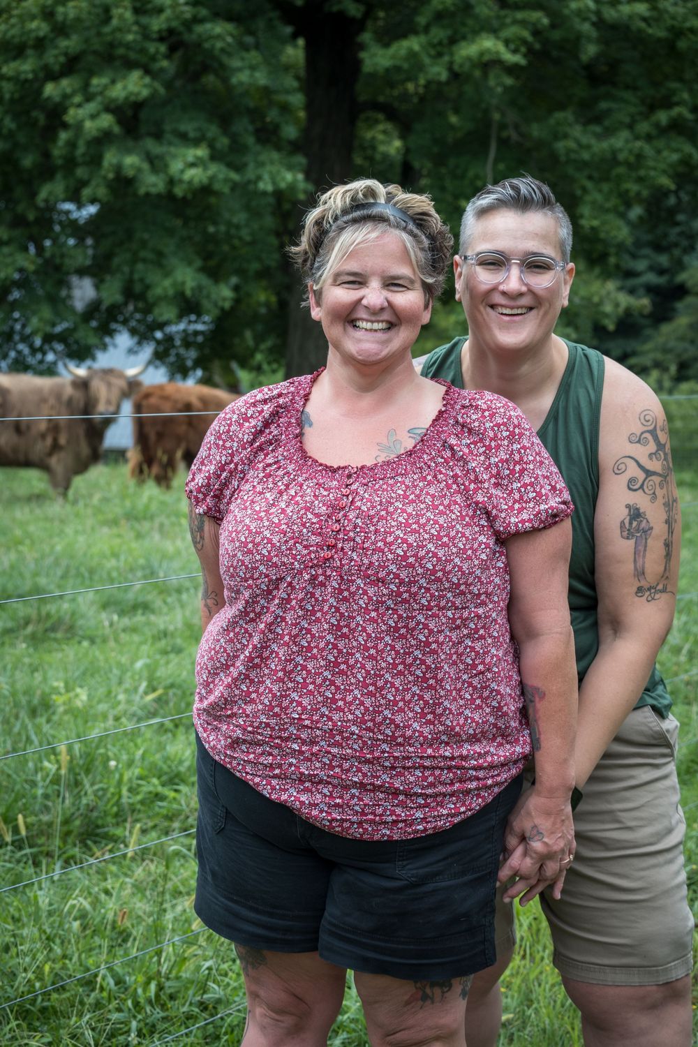 Two people in casual summer clothing smile outdoors with green trees and grass in the background.