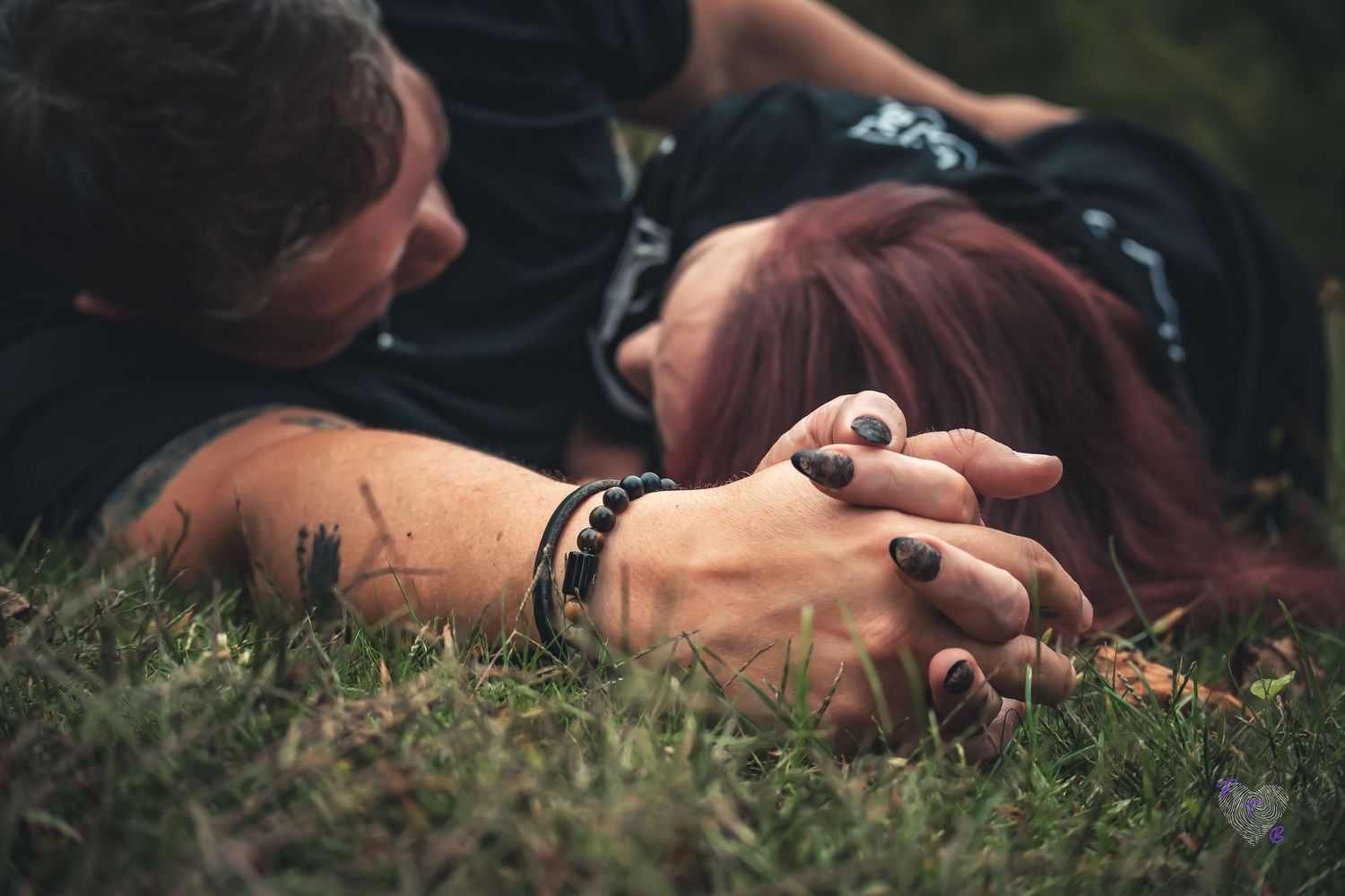 Couple lying on grass holding hands tenderly, woman with red hair and dark nail polish, romantic outdoor moment.
