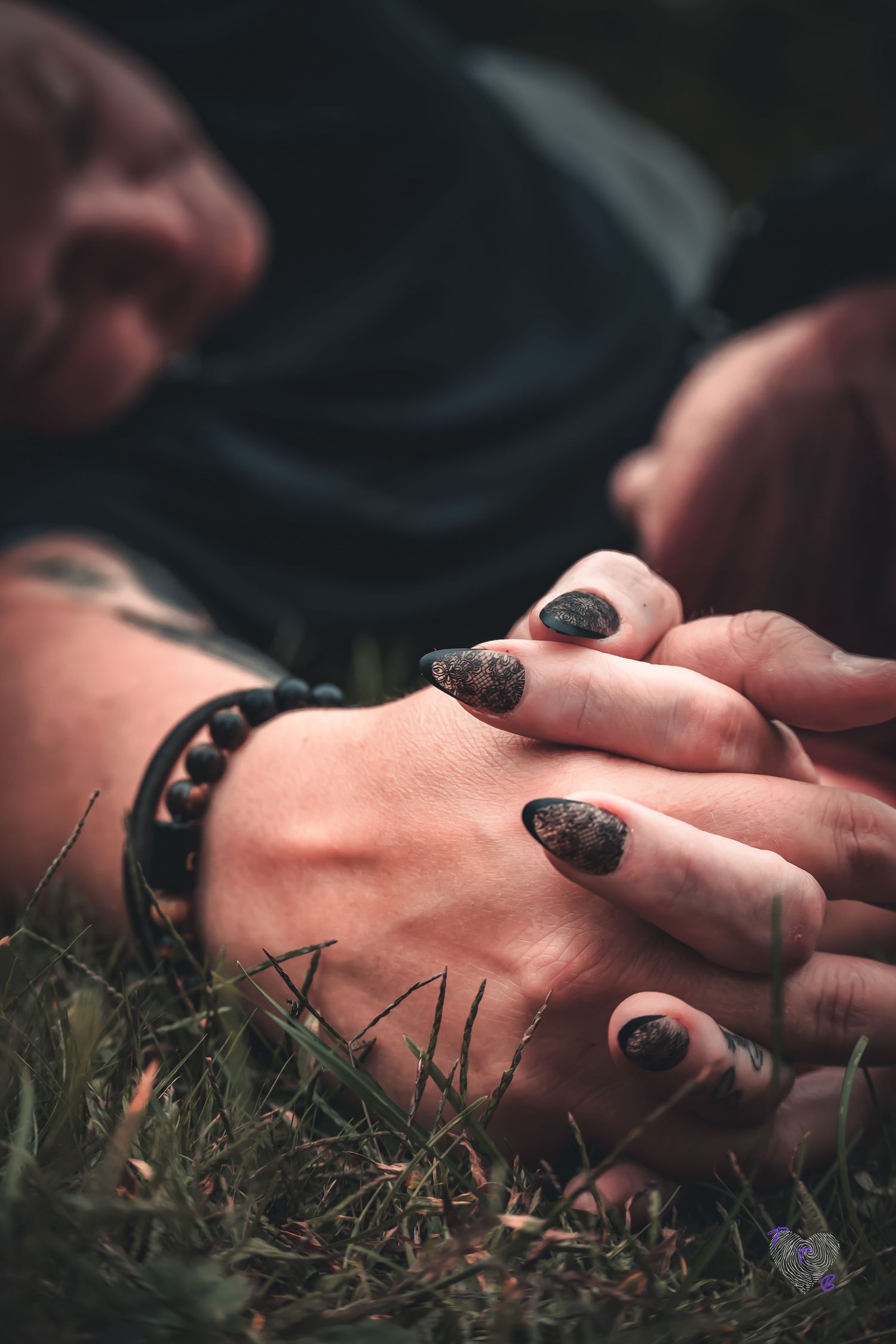 Two hands intertwined on grass, one with dark patterned nails and a beaded bracelet, with a blurred person in background.