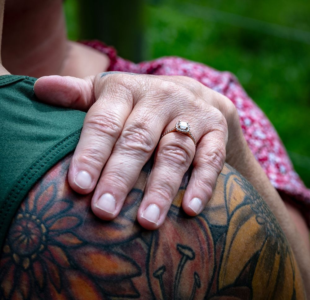 A hand wearing an engagement ring rests gently on a tattooed body with floral designs and vibrant colors.