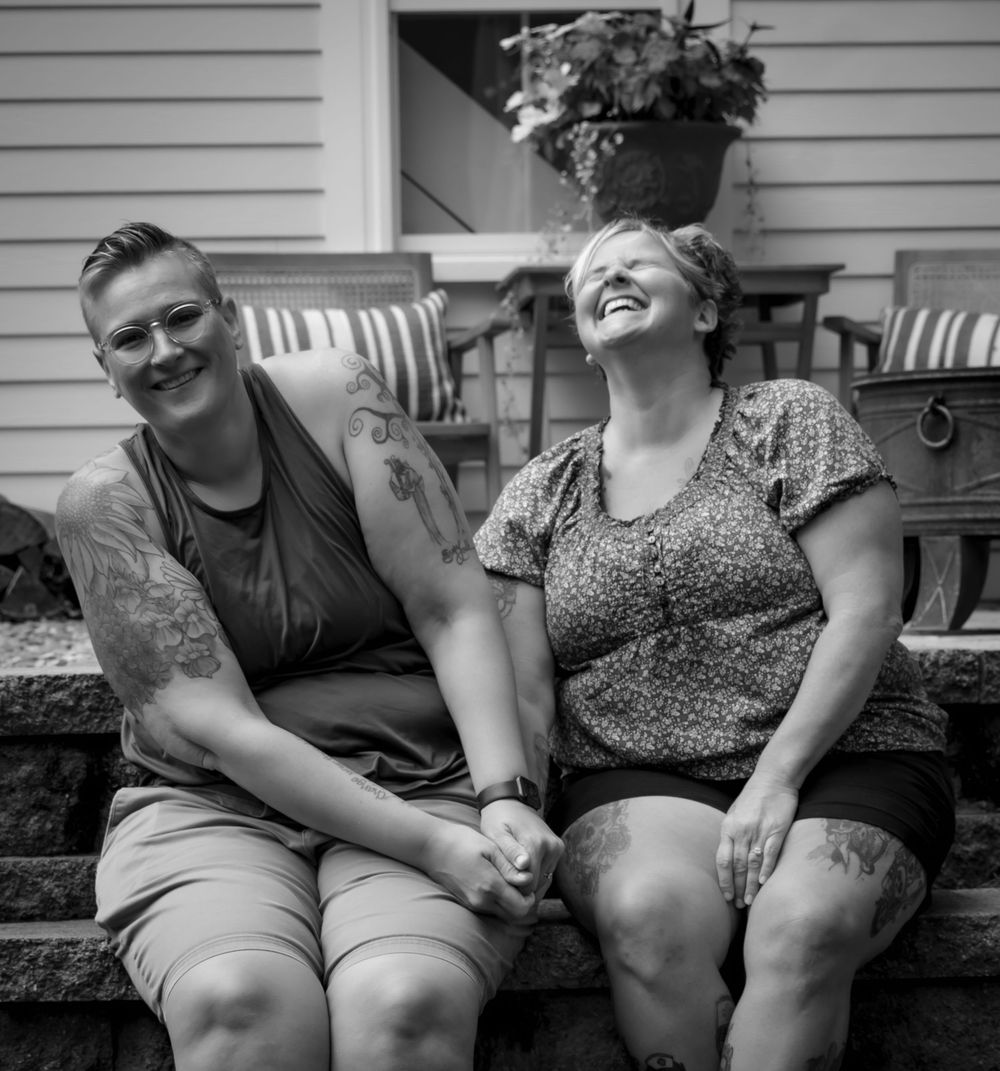 A couple sitting closely together on a porch share a joyful moment of genuine laughter in black and white.