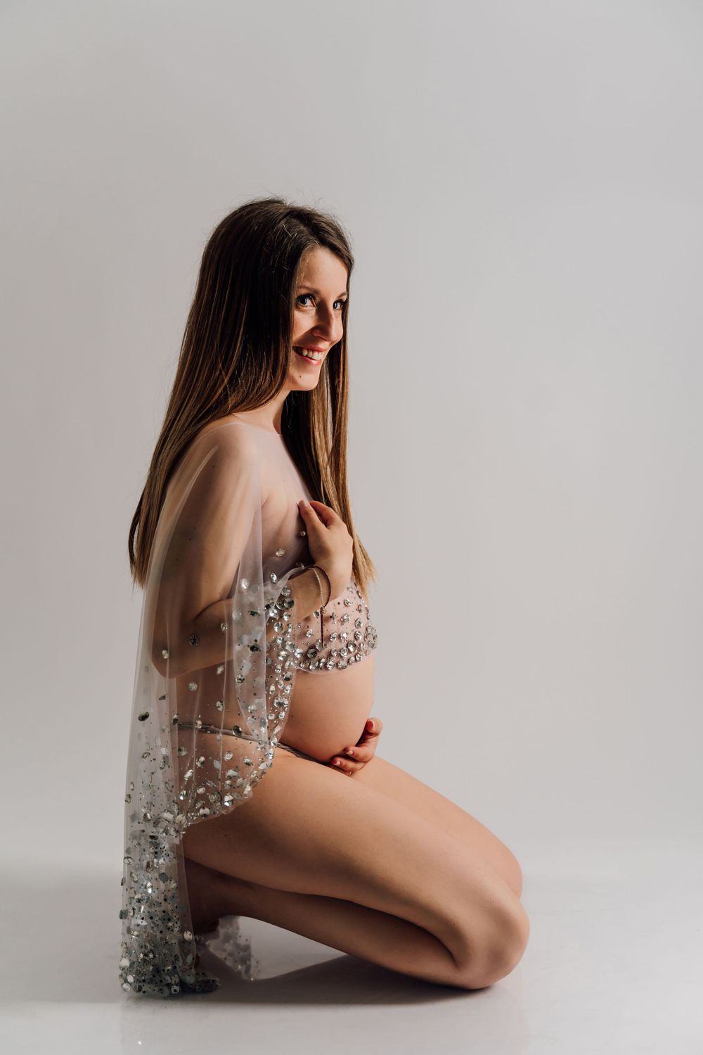 Ethereal maternity pose in sheer sequined robe against light background.