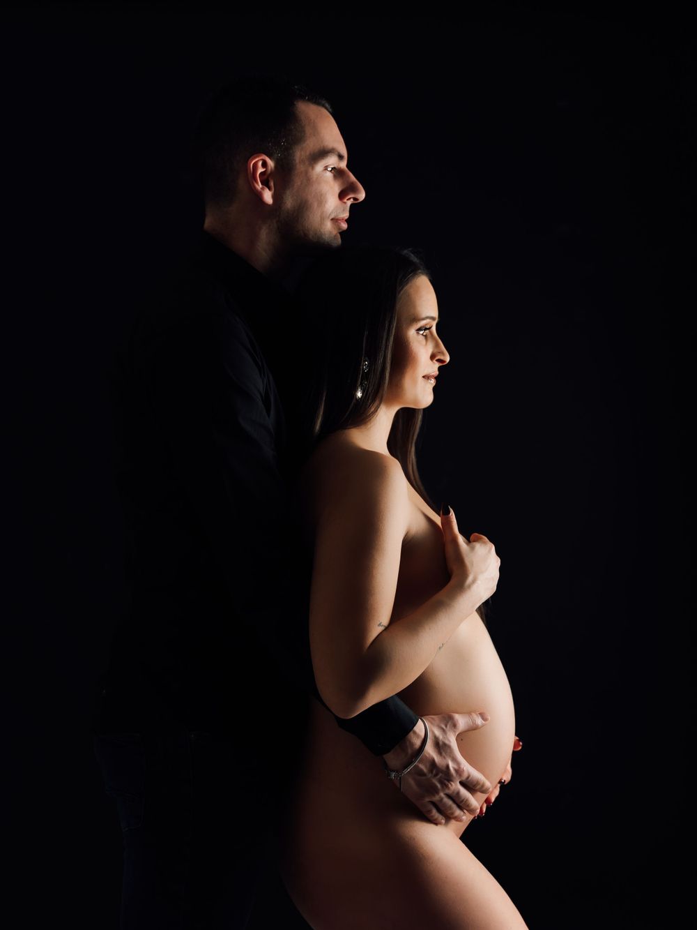 Artistic maternity photo silhouette against dark background showing couple in intimate moment.