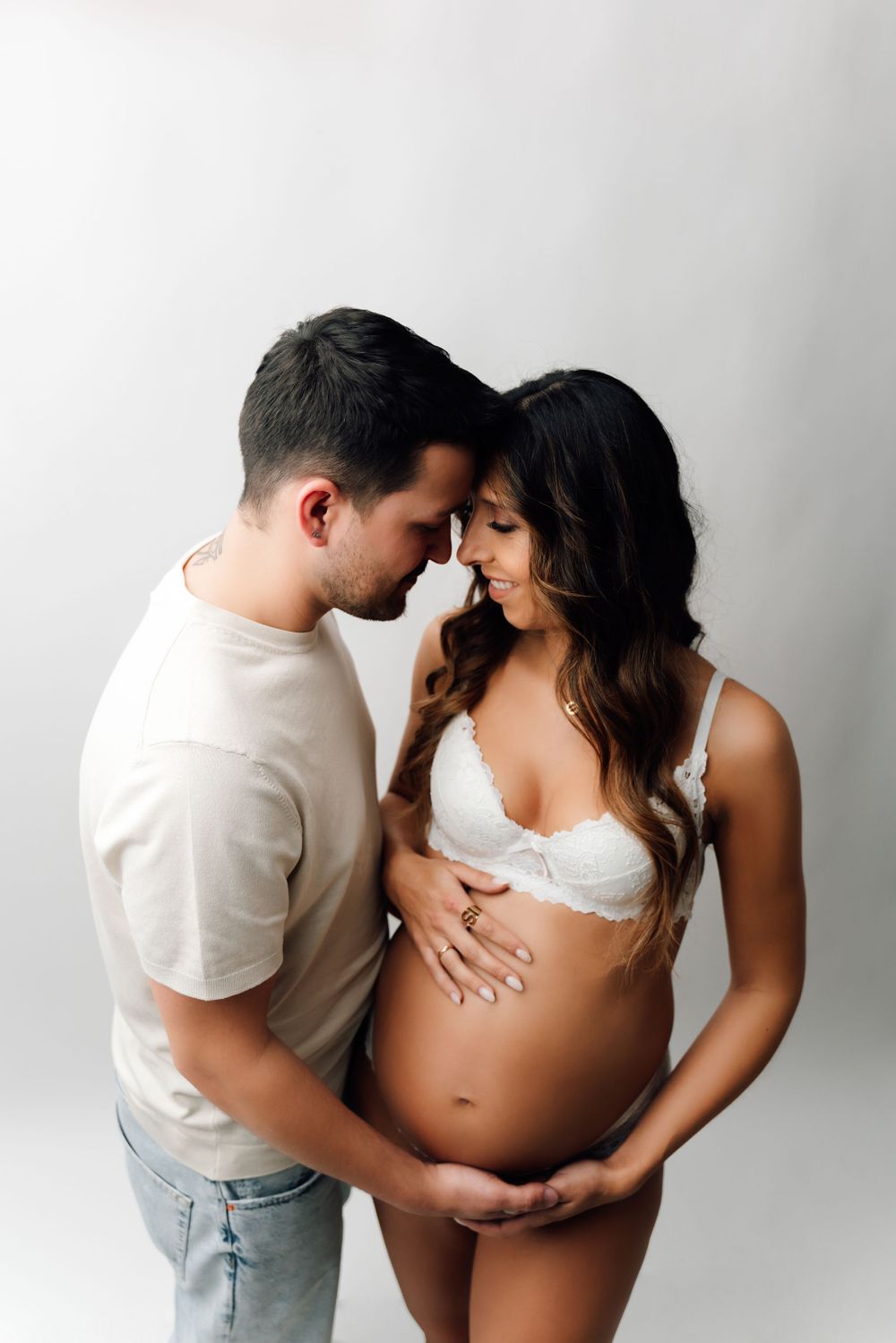 Intimate maternity portrait of couple in white clothing against light background.