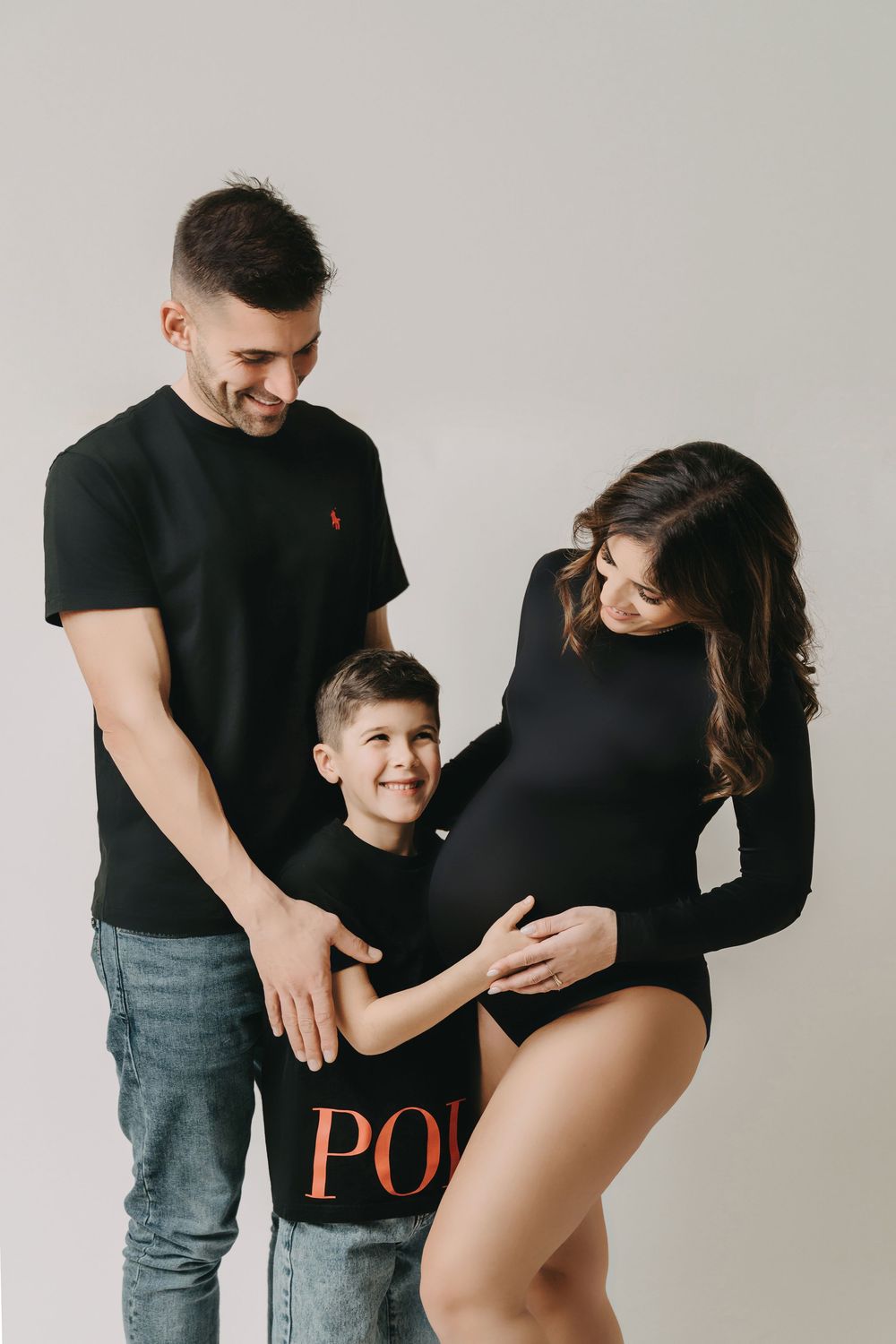 Family maternity photo in studio showing parents in black outfits with young child touching mothers pregnant belly.
