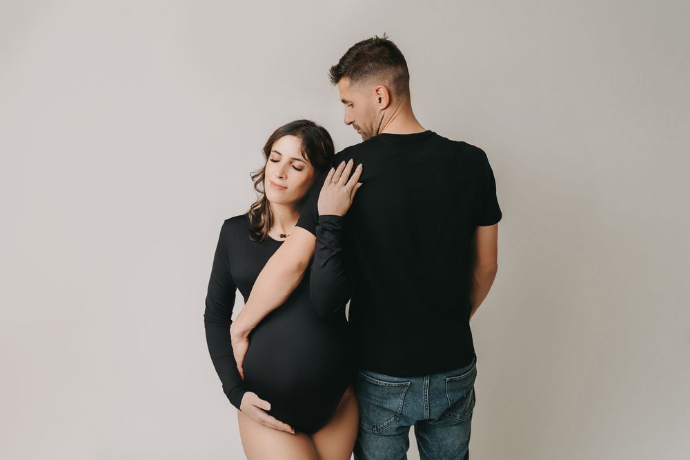 A couple poses intimately against a neutral wall backdrop during a minimalist maternity photoshoot.