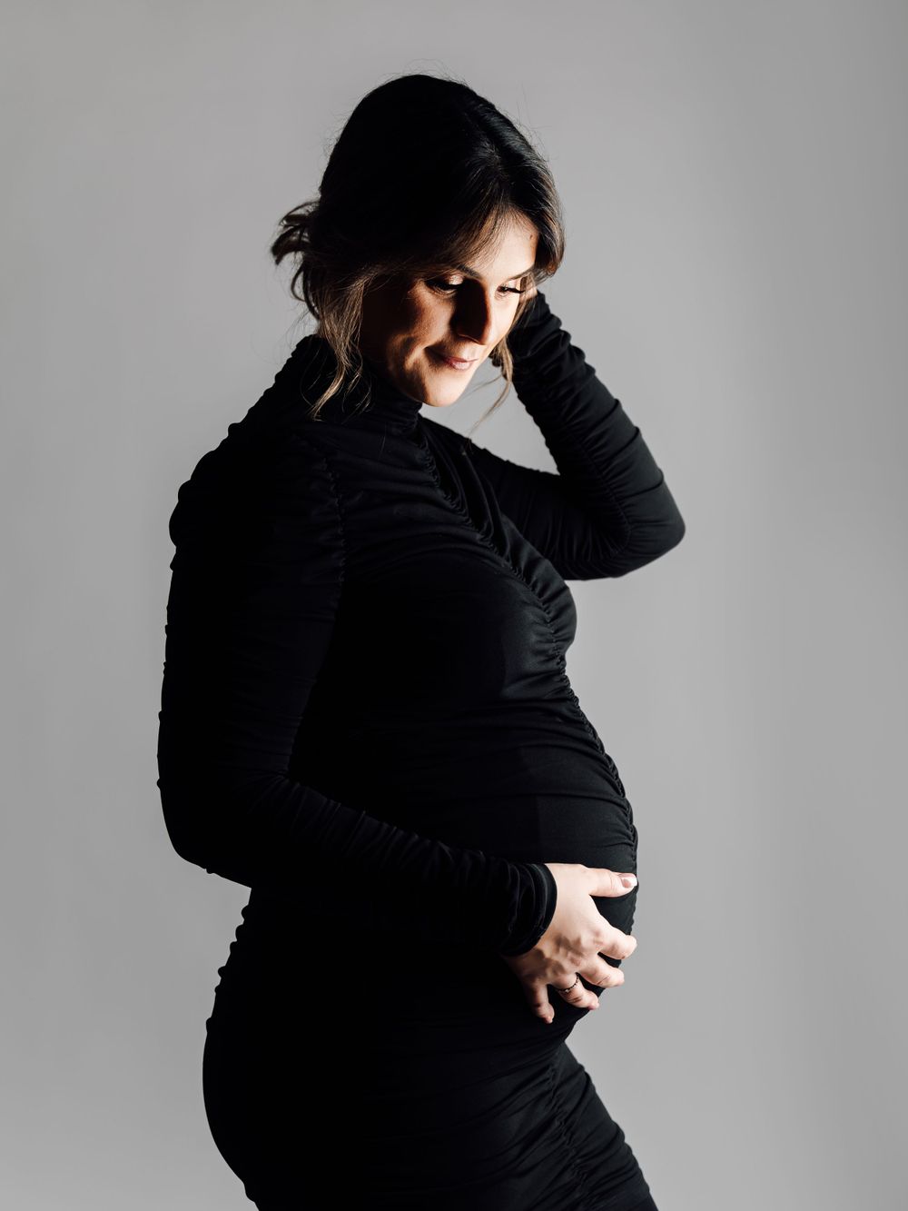 Elegant silhouette of pregnant woman in black dress posing against light grey studio backdrop.