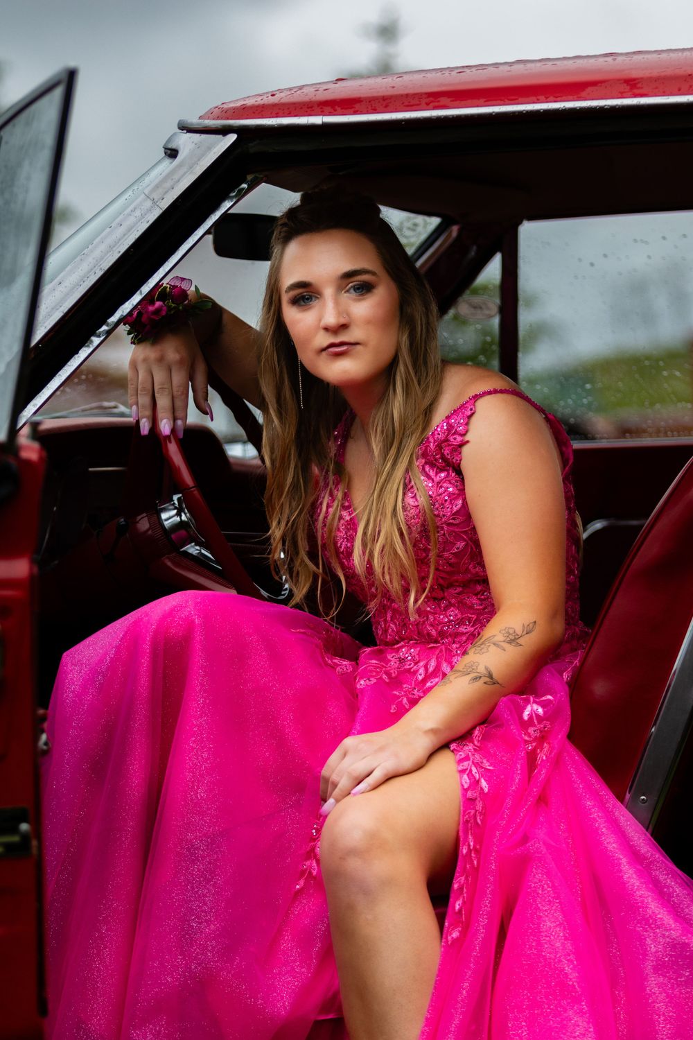 Pink formal gown contrasts with vintage red car interior in stylish portrait setting.