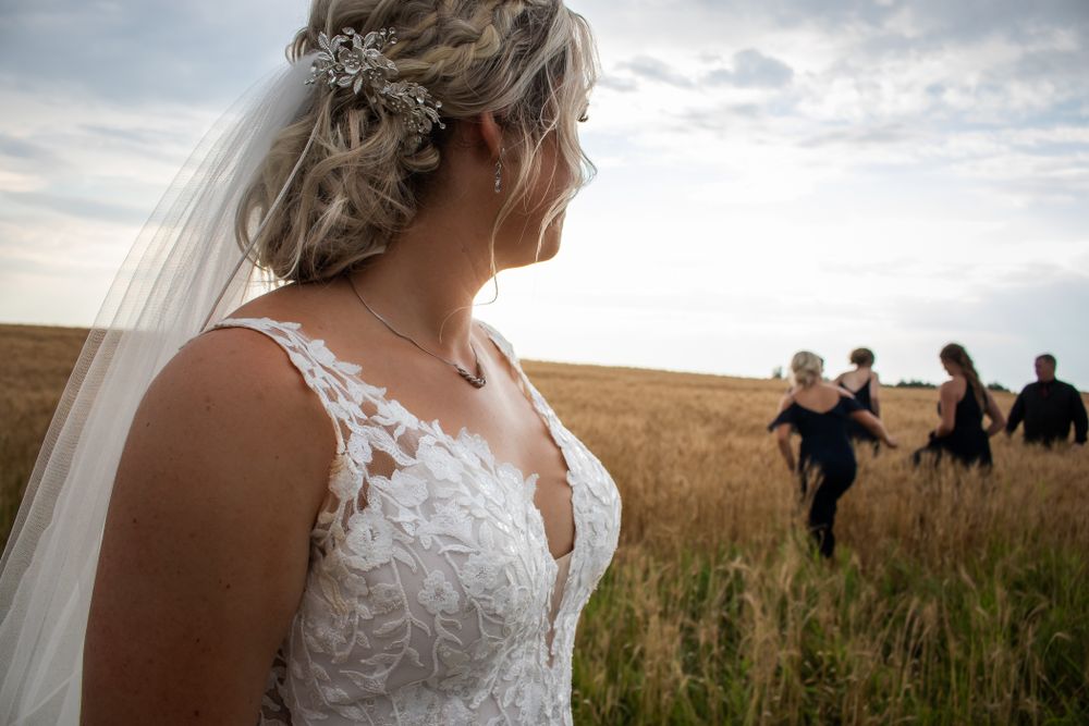 A bride in a lace dress stands in a wheat field while others celebrate in the background.