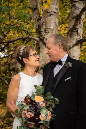 A couple shares an intimate moment under autumn birch trees with a peach and burgundy bouquet.