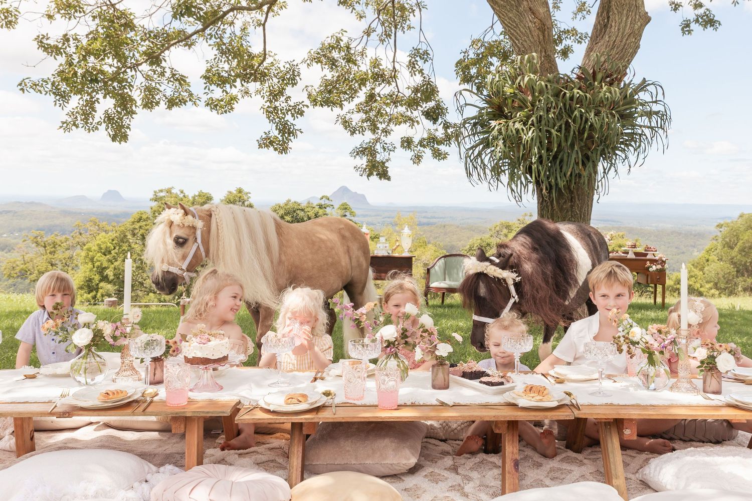 Elegant outdoor tea party setting with pink decor and horses grazing in the background.