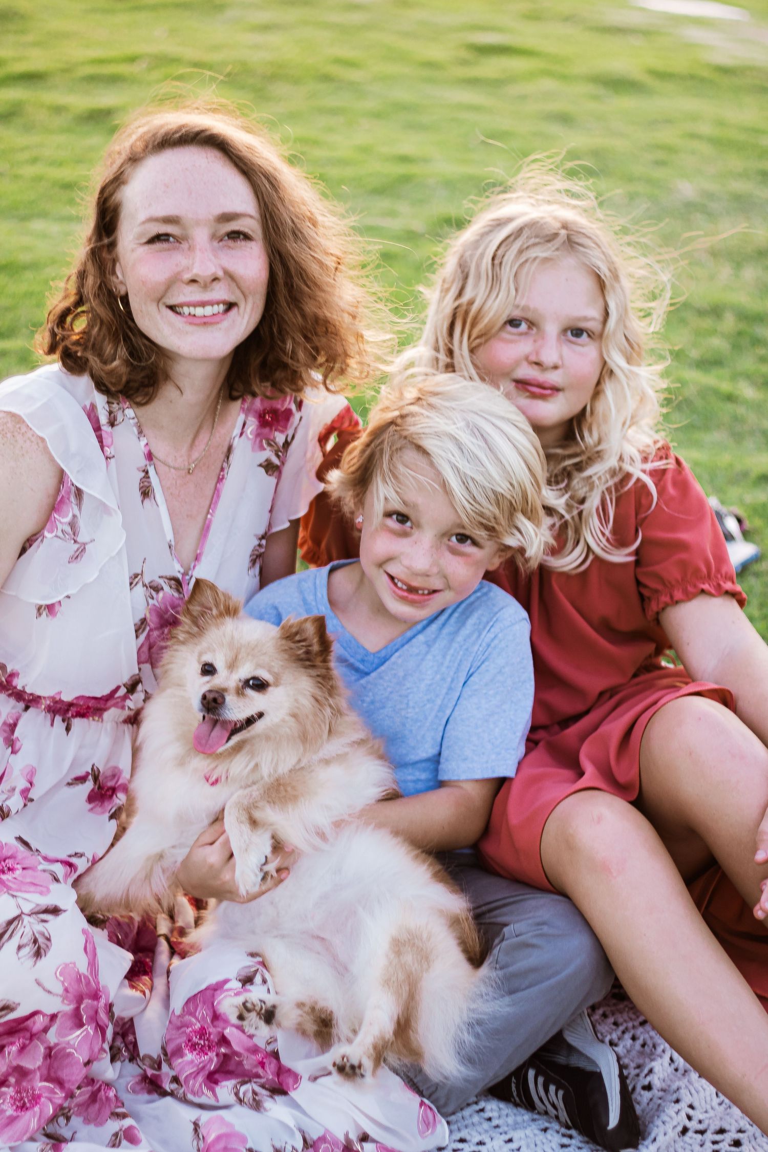 Happy family relaxes on grass with small fluffy dog while wearing floral dresses.