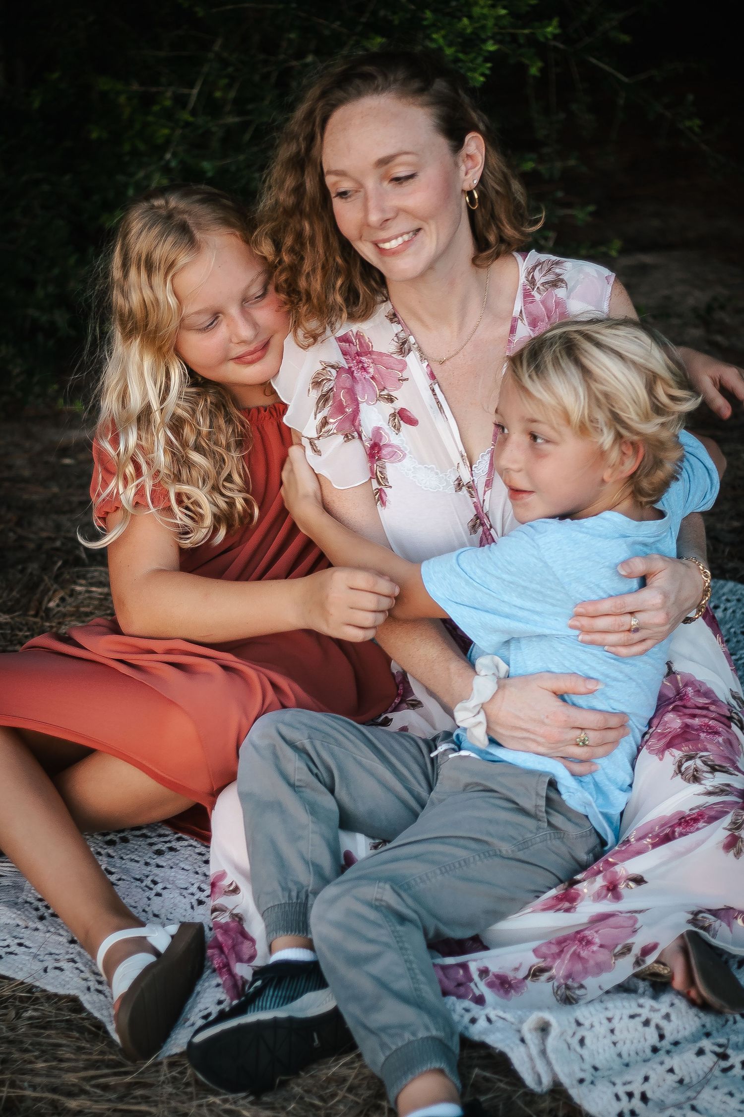 Family cuddles together on outdoor bench wearing floral and casual clothing.
