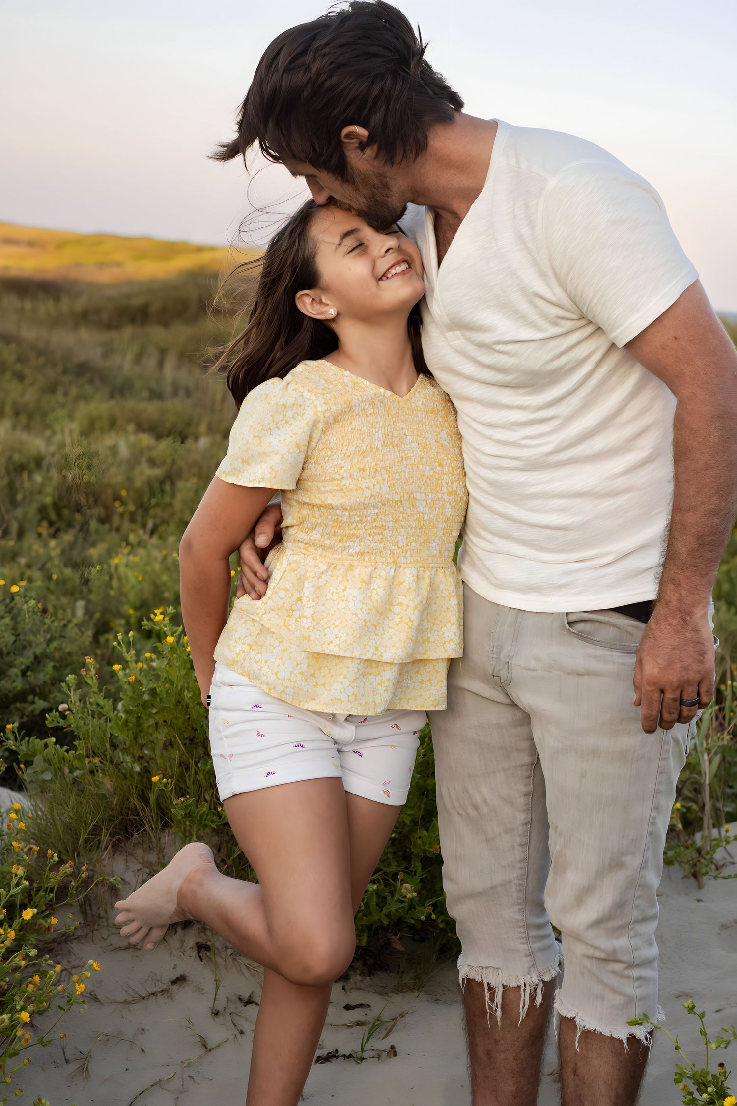 Family in casual summer outfits shares playful moments together on sandy beach.