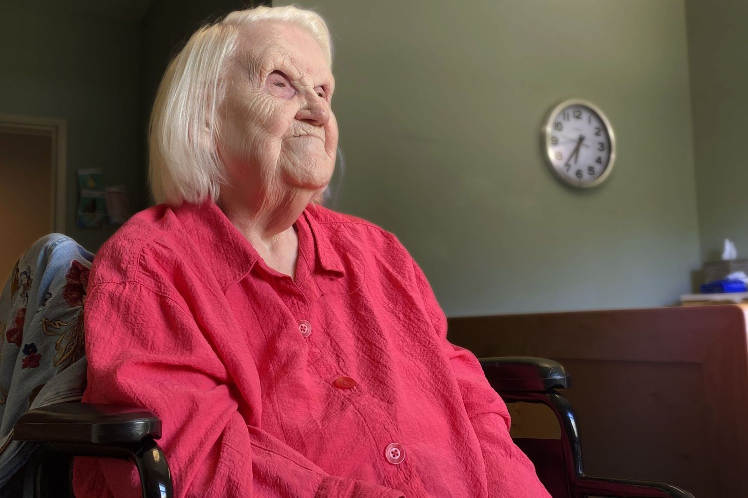 Elderly woman sits in a wheel chair with white straight hair and a slight smile. The woman is Maxine Kent