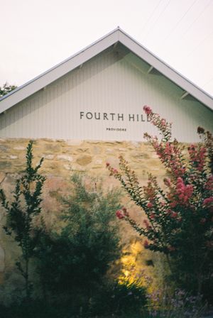 White rustic building with Fourth Hill signage against mountain backdrop and colorful landscaping.
