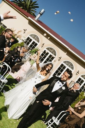 Joyful newlyweds walk through confetti shower after outdoor wedding ceremony, guests cheering beside white venue building.