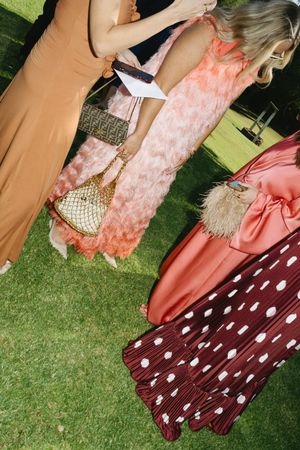 Women in elegant pink and orange gowns holding designer bags at an outdoor garden party event.