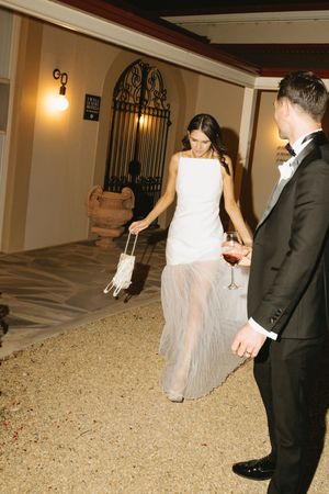 Bride in white gown with sheer skirt holds purse beside groom in tuxedo holding wine glass at evening venue.