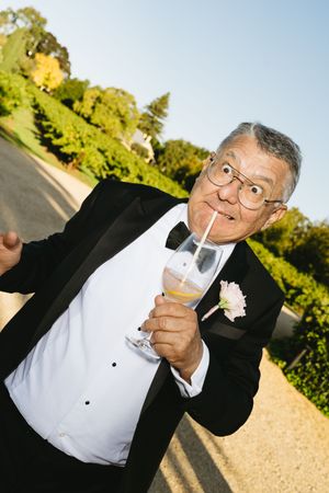 Elderly man in tuxedo with bow tie sipping champagne through straw at outdoor wedding celebration.