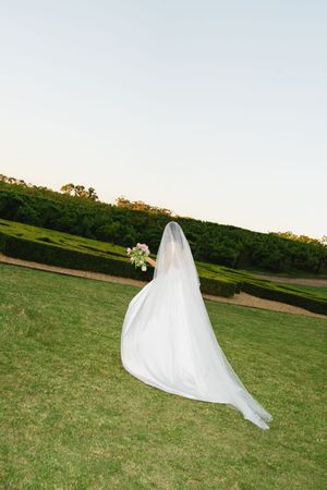 Bride in flowing white gown and veil holding bouquet stands on lush green lawn at elegant outdoor wedding venue.
