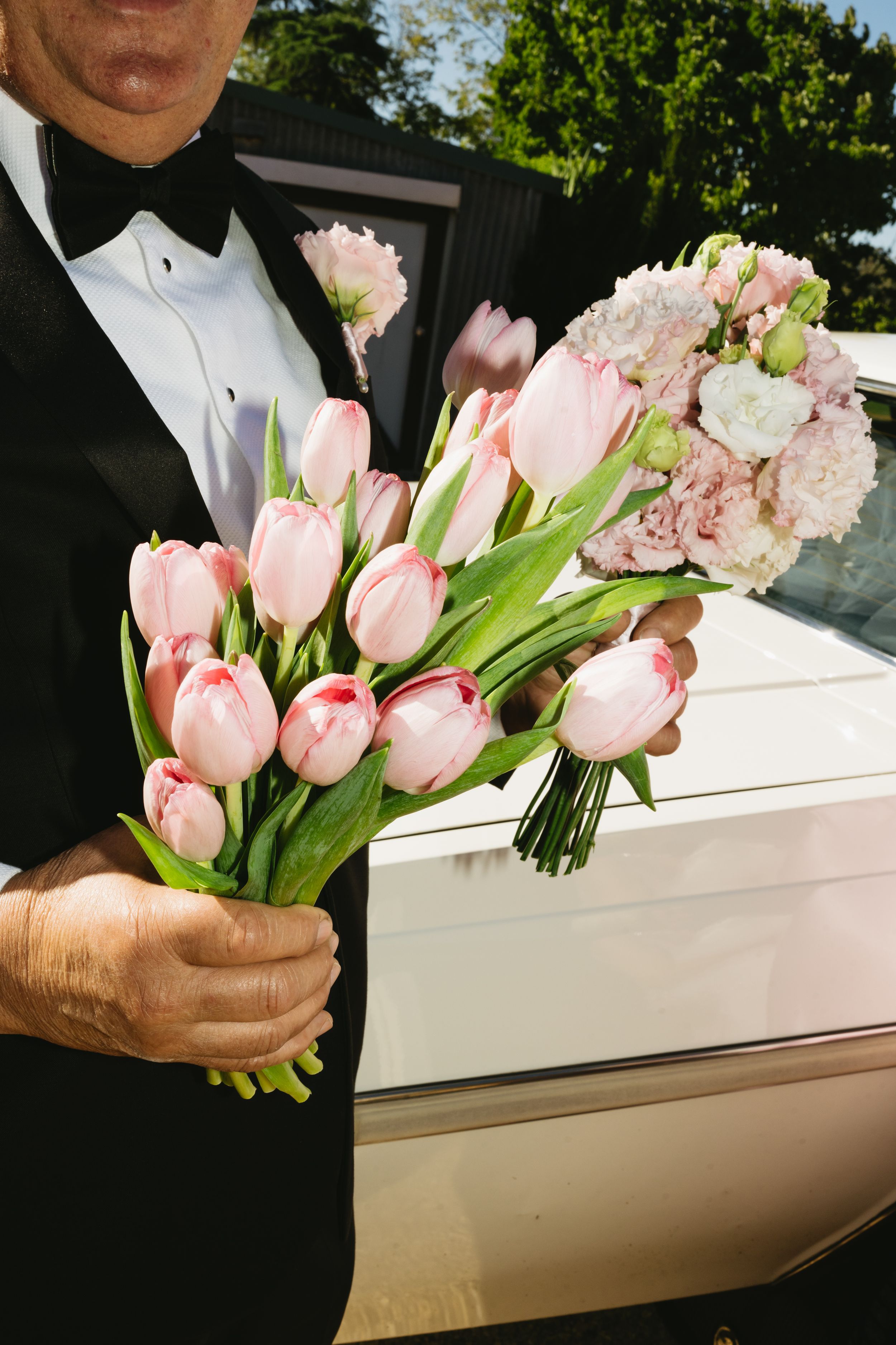 Man in tuxedo holding two pink tulip and carnation wedding bouquets beside a white limousine outdoors.