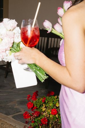 Woman in pink dress holding a red cocktail with straw and floral bouquet at an outdoor wedding event.
