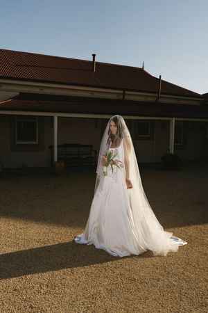 Bride in white gown and veil holds bouquet outdoors before rustic building on gravel ground under clear blue sky.