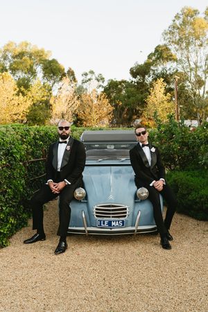 Two grooms in black tuxedos pose beside a vintage blue Citroen 2CV car on gravel, surrounded by lush hedges.