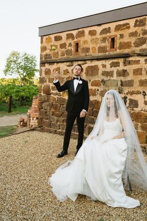 Bride in white gown and veil sits while groom in tuxedo tosses object near rustic stone building.
