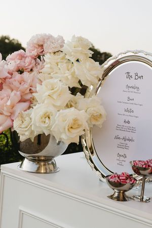 Elegant wedding bar display with white roses, pink peonies in silver vase, framed bar menu, and candy dish on white cabinet.