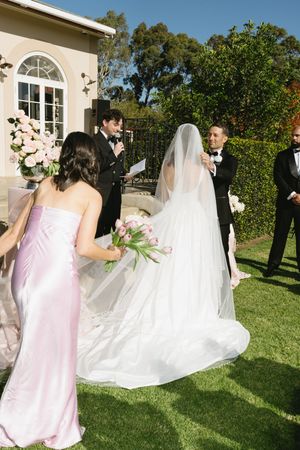 Outdoor wedding ceremony with bride in white gown and veil, groom, officiant, and bridesmaid in pink dress on green lawn.
