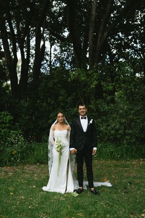 A couple in wedding attire stands together on a grassy lawn surrounded by trees in an outdoor garden setting.