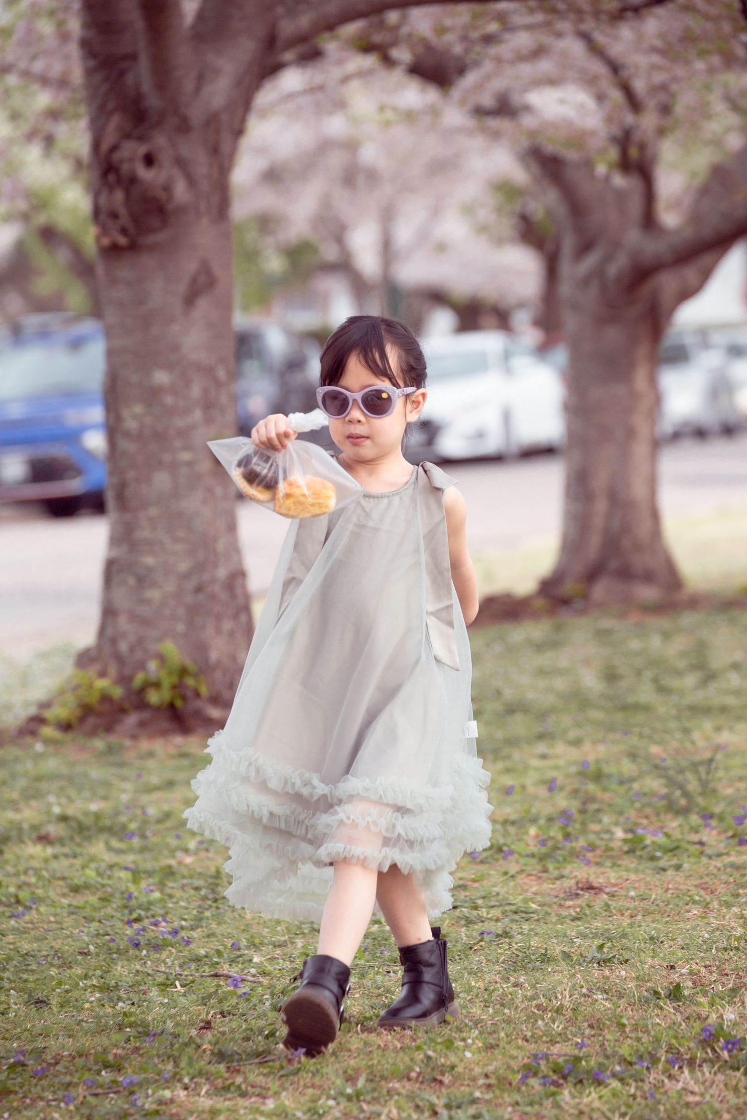 Child in ruffled dress and boots walking in park holding snack.