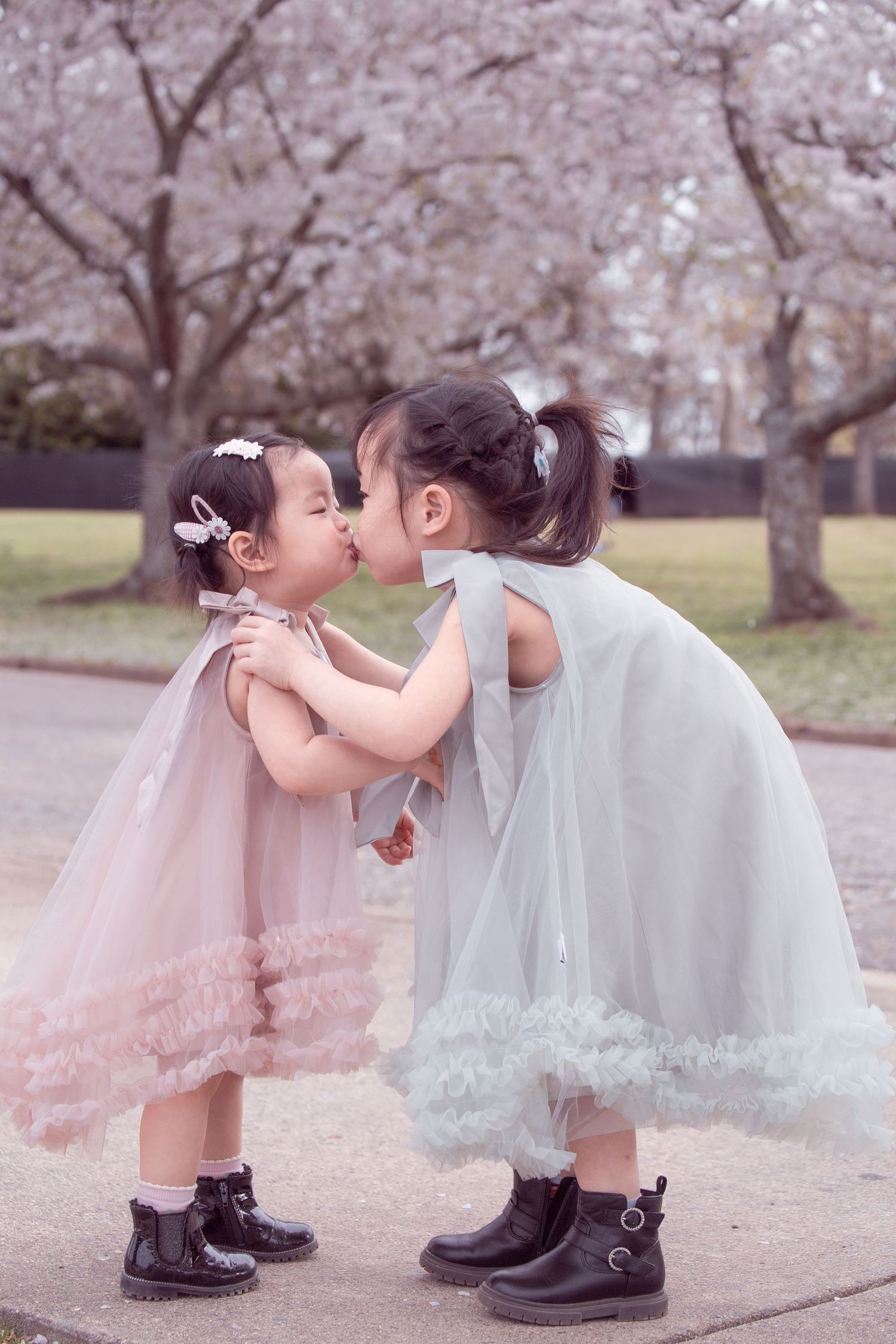 Two children in matching dresses and boots sharing sweet moment under cherry blossoms.