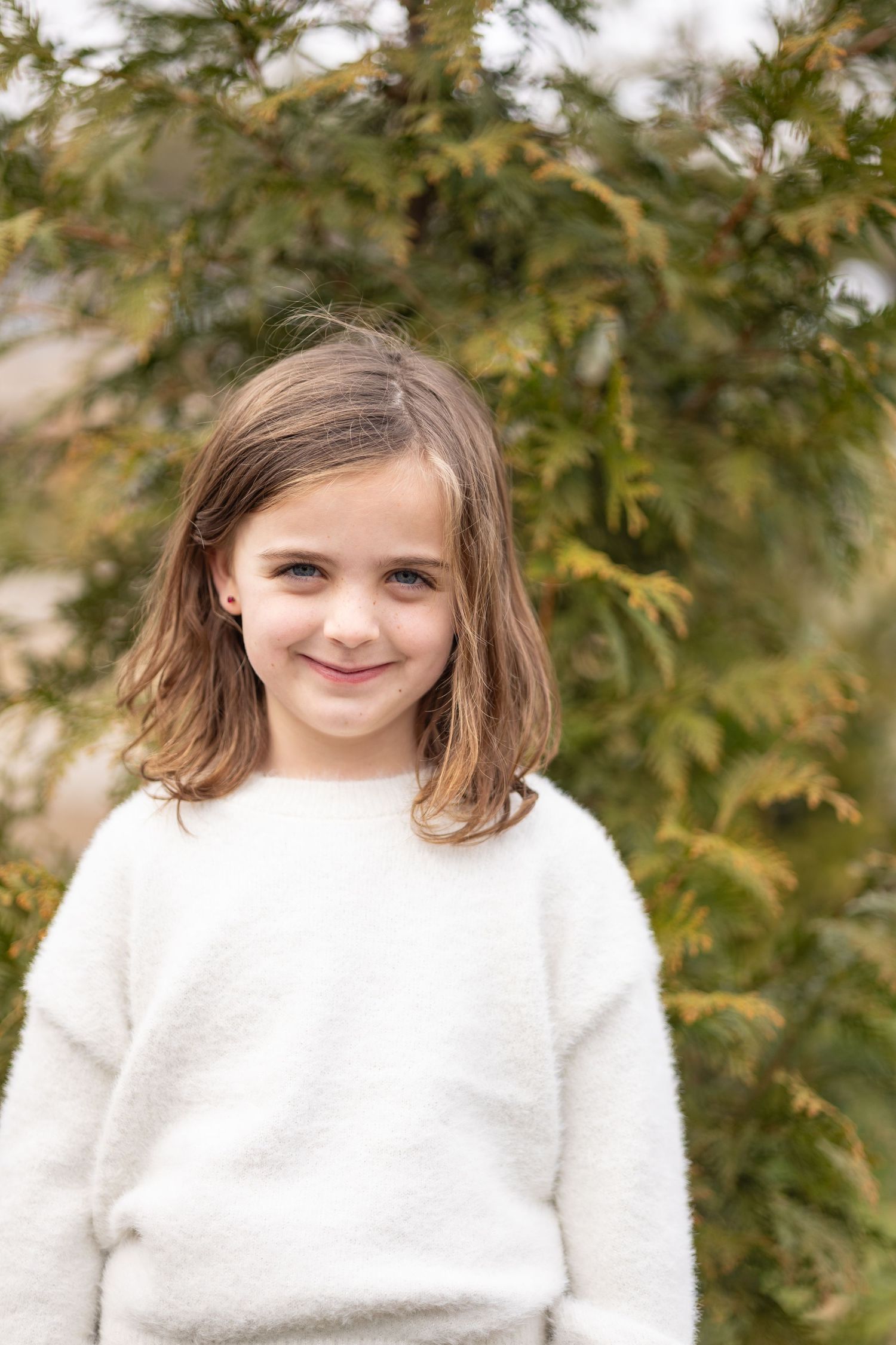 A child wearing a cozy white sweater stands in front of green foliage on a sunny day.