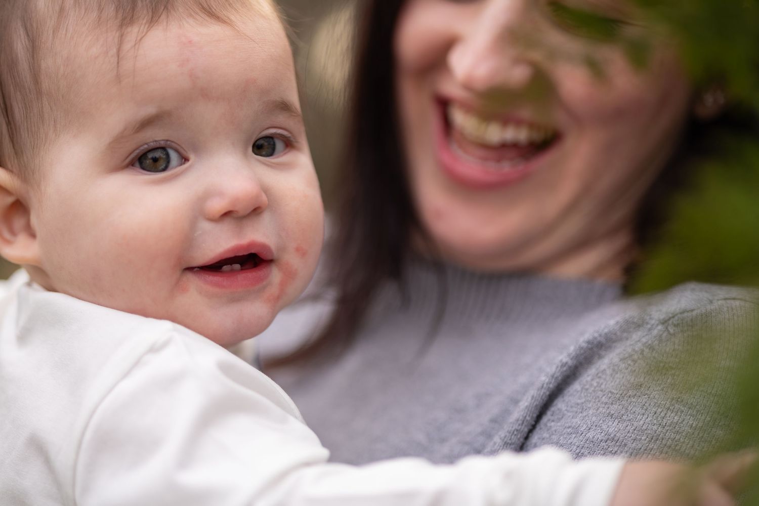 A baby smiles and plays while sitting on someone's lap outdoors in a garden setting.