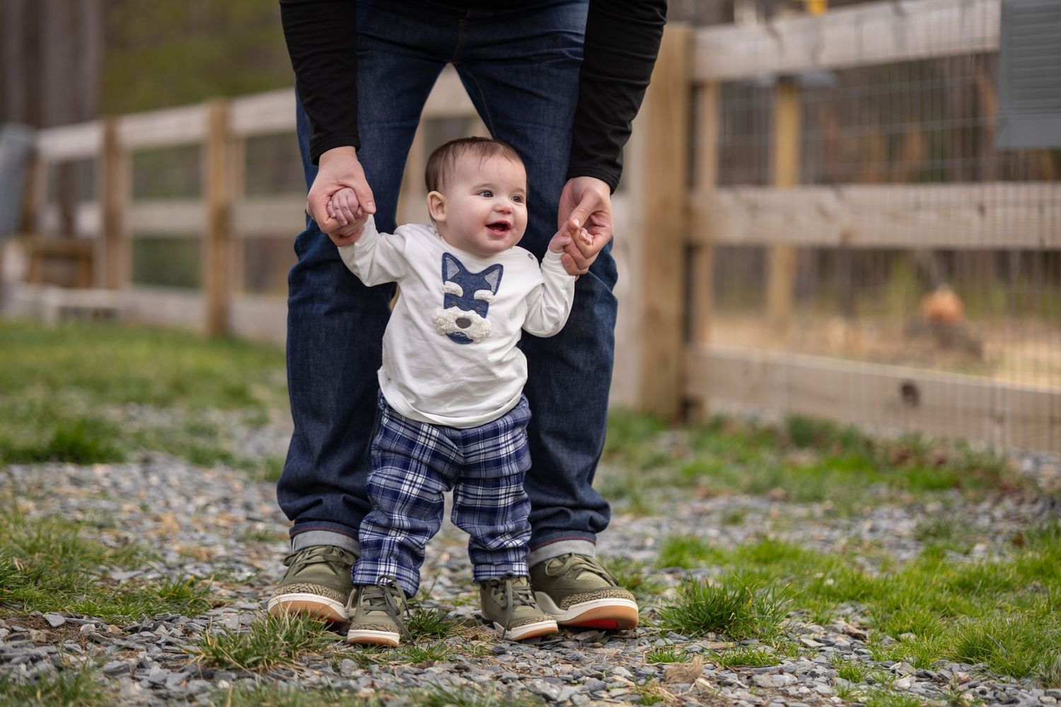 Adult in blue jeans holds a small child's hands while helping them walk on a gravel path outdoors.