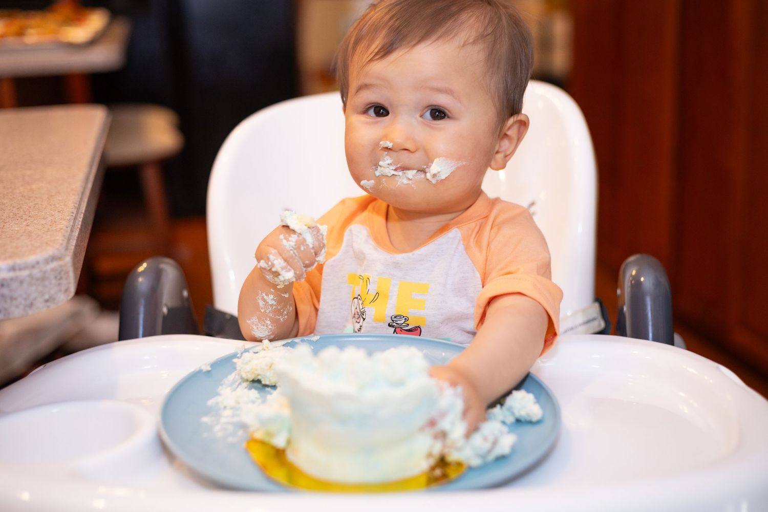 Cute baby in orange bib smashing first birthday cake in high chair, face covered in white frosting.