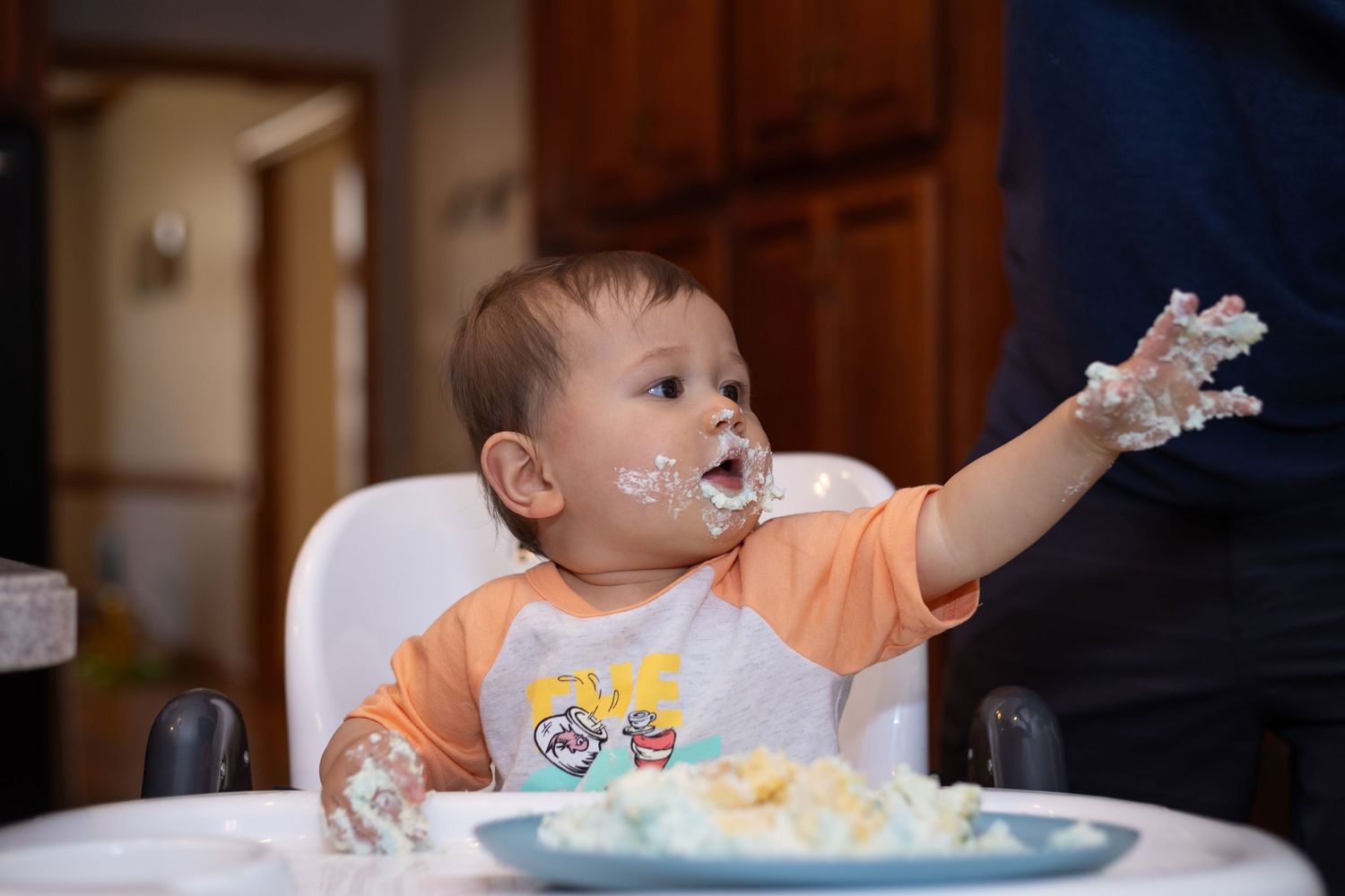 Messy baby in high chair with cake frosting on face and hand during first birthday celebration.