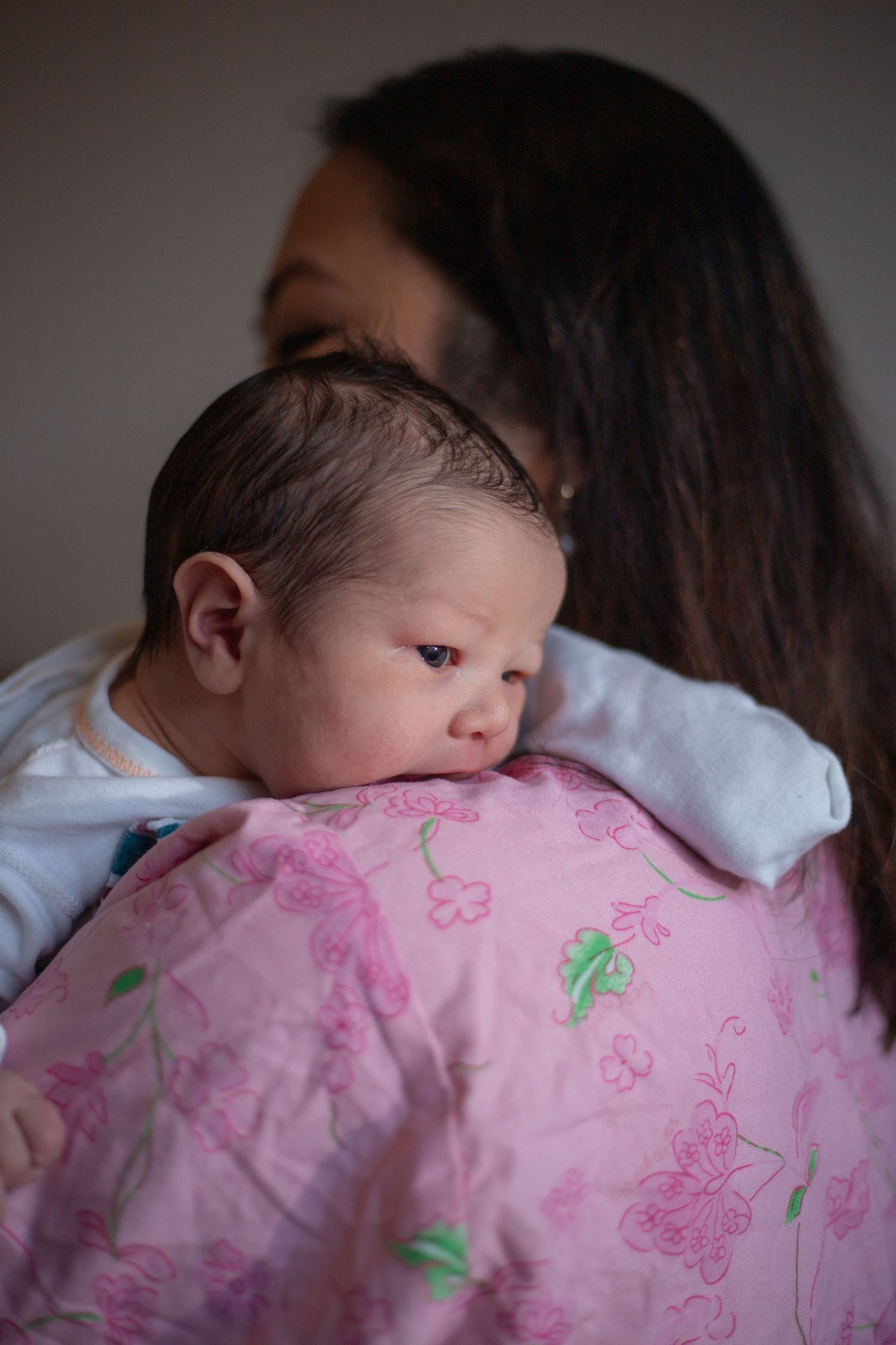 A tiny infant rests against a pink floral blanket while being held by someone with long dark hair.