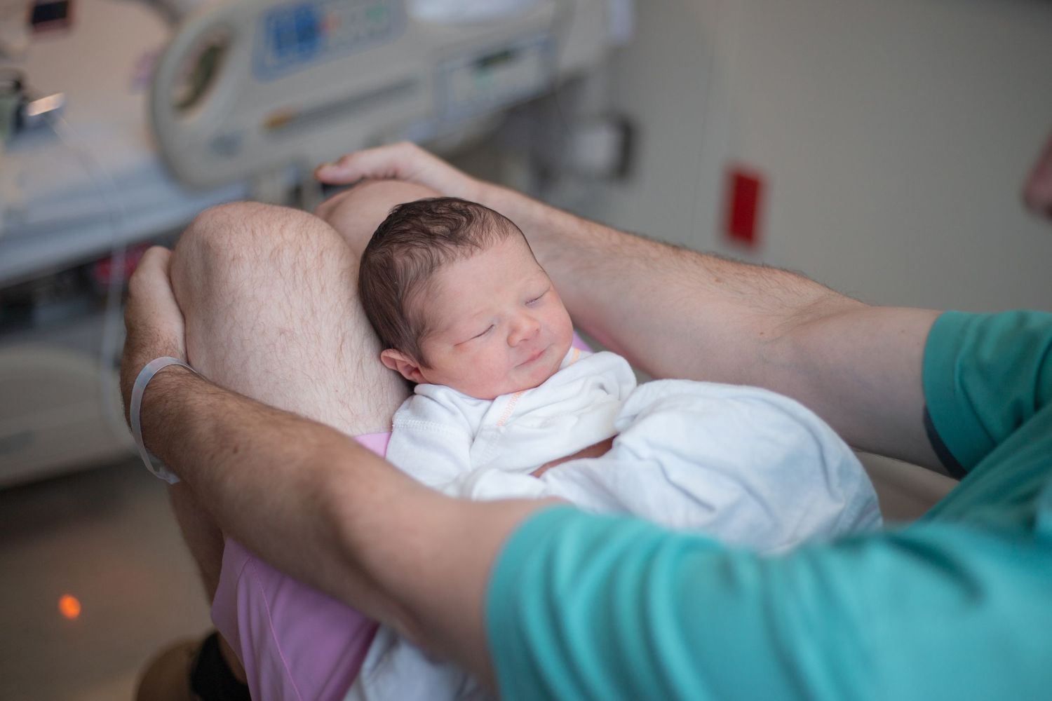 A sleeping newborn wrapped in white cloth is gently cradled in hands wearing turquoise medical sleeves.