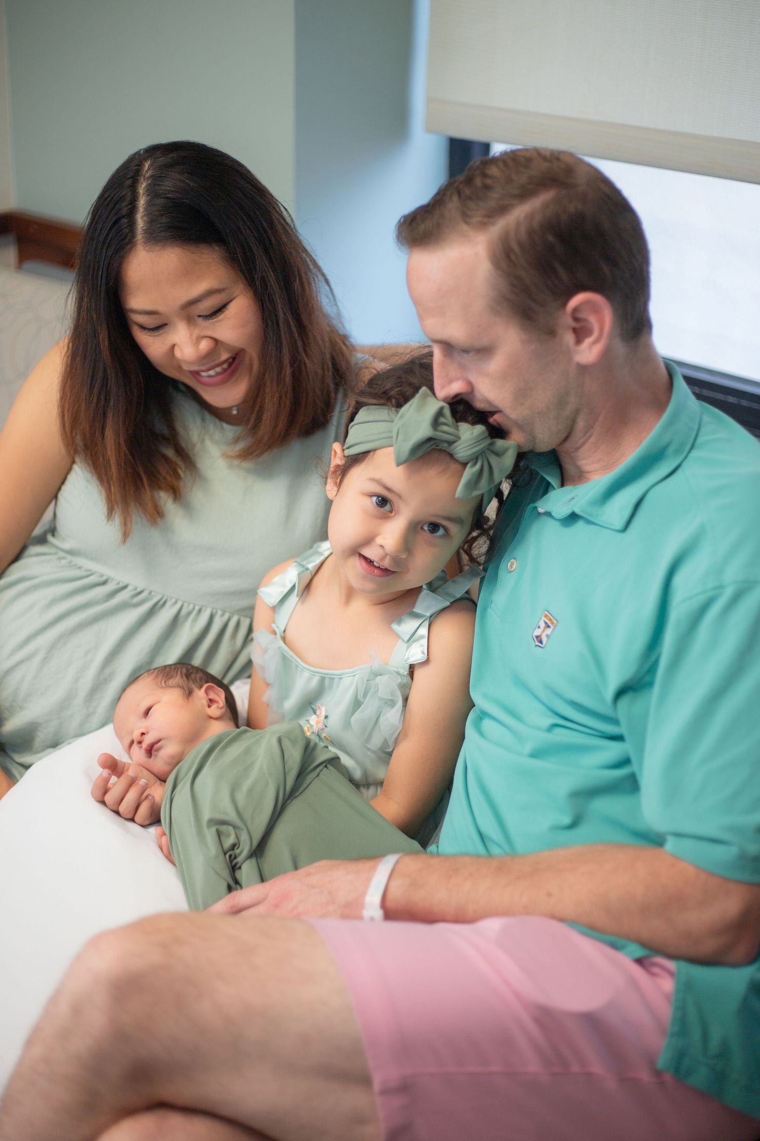A family sits together on a couch with their newborn baby and young daughter in matching sage green outfits.