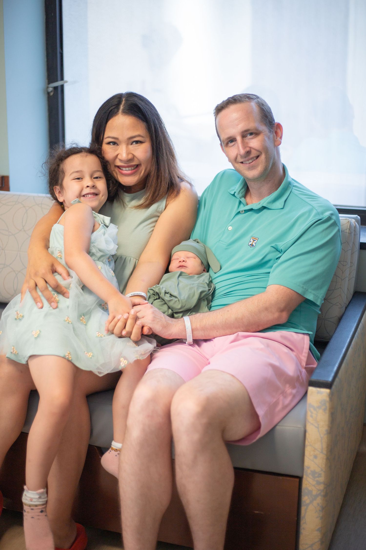 A happy family relaxes together on a couch, with one adult wearing a sage green top and another in a mint polo shirt.