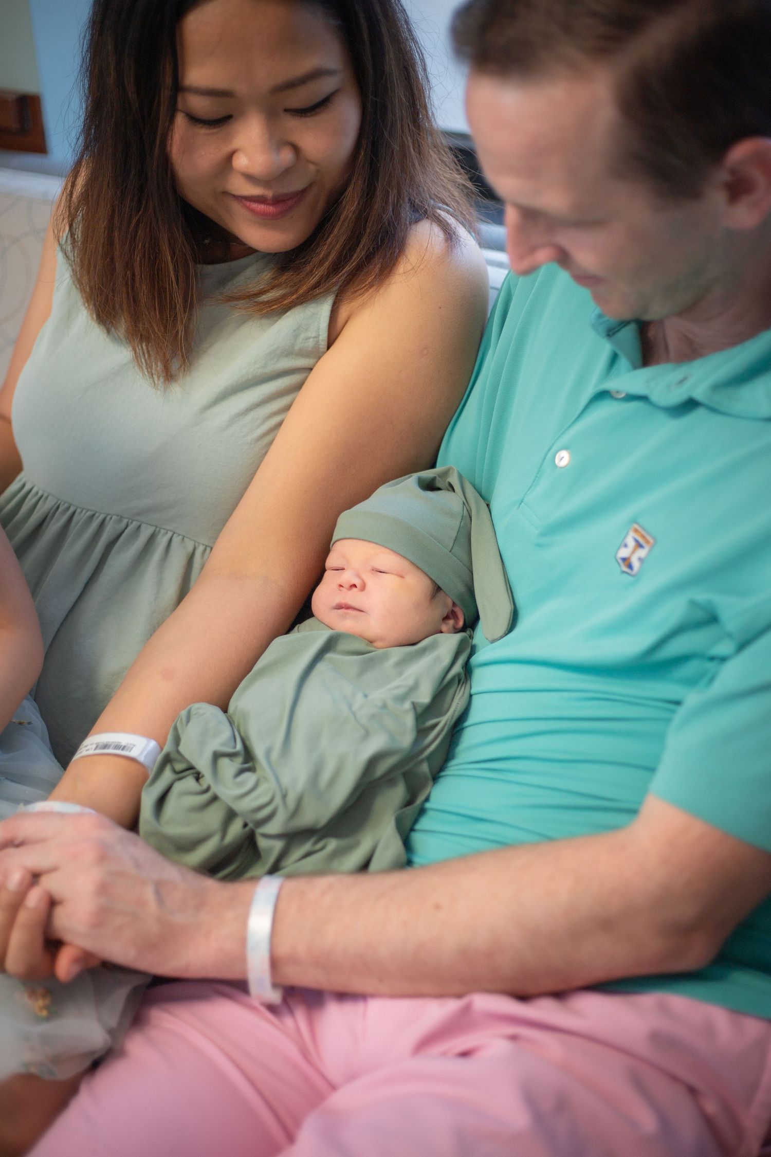 A couple gazes lovingly at their sleeping newborn baby wearing a sage green outfit and hospital bracelet.