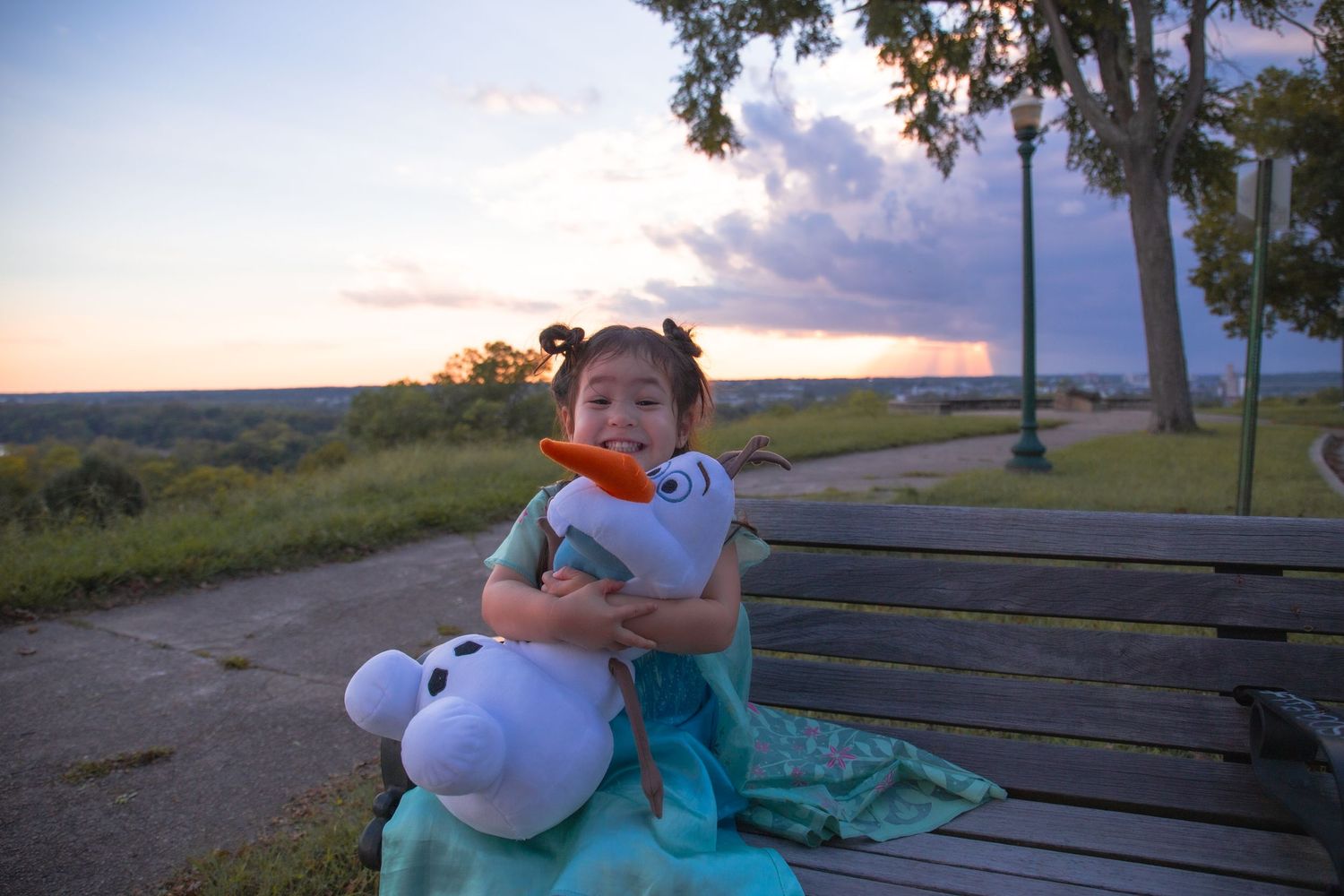Child sits on park bench at sunset with stuffed animal toy.