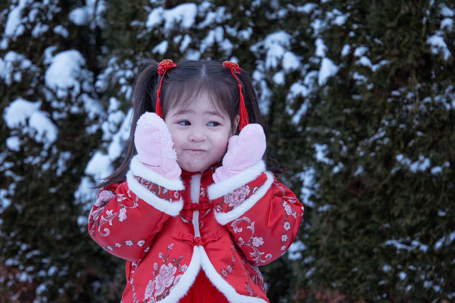 Child wearing red Chinese dress and white mittens poses against snowy background.